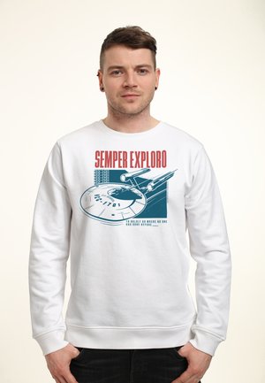 Mann trägt weißen Pullover mit Raumschiff-Grafik und dem Text „Semper Exploro“ und „Kühn dorthin gehen, wo noch niemand war.“