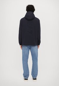 Mørkeblå hettejakke med et preget "EA"-logo på baksiden, kombinert med lyseblå jeans og svarte joggesko.
