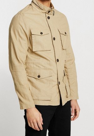 Mężczyzna w beżowej zapinanej na guziki kurtce z czterema przednimi kieszeniami na klapki i czarnych spodniach, stojący na jednolitym tle.