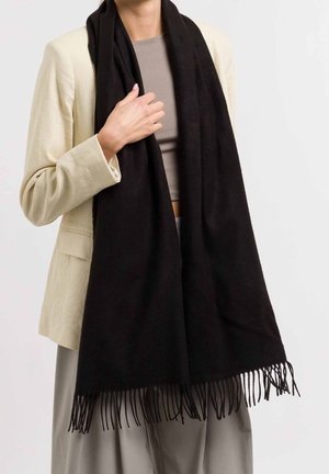 Personne portant un blazer beige et un haut gris, tenant une longue écharpe noire avec des extrémités à franges drapée autour du cou.