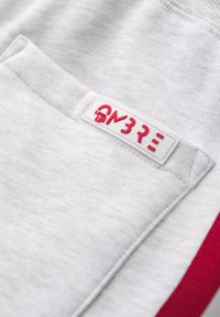 Material de sudadera gris con una superficie texturizada, que presenta una etiqueta blanca con el texto rojo "ON3RE LEVEL UP" en el bolsillo. Acento rojo a lo largo del costado.