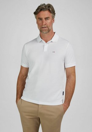 Mann mit grauem Haar und Bart, bekleidet mit einem weißen Poloshirt und beigen Hosen, steht mit den Händen in den Taschen vor einem schlichten Hintergrund.