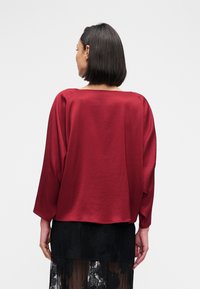 Blouse en satin bordeaux à coupe décontractée, manches larges et encolure carrée, associée à une jupe en dentelle noire transparente.
