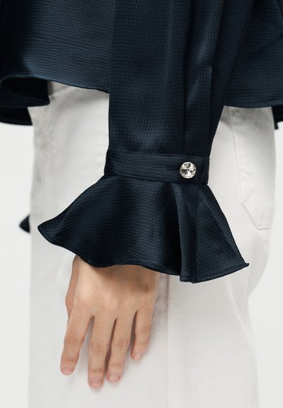 Χέρι ακουμπισμένο δίπλα σε λευκό παντελόνι, φοράει μαύρο υφασμάτινο μανίκι με φουσκωτό μανσέτα και ένα διαφανές κουμπί ως λεπτομέρεια.