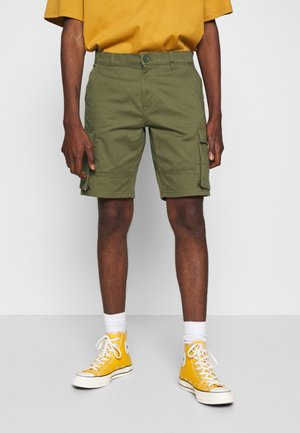 Personne portant un short cargo vert olive, des chaussettes blanches et des baskets montantes jaunes, debout devant un fond uni.
