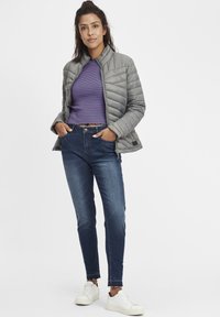 Pelēka izolēta jaka ar augstu apkakli, vilkta pār svītrainu zilas un violetas krāsas īsu topu, kopā ar zilām džinsa biksēm un baltām sporta kurpēm.