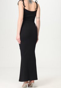 Abito maxi nero a coste con spalline regolabili, design aderente e orlo dritto, con un tatuaggio sottile visibile sulla schiena della indossatrice.