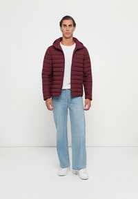 Mand står iført en kastanjebrun pufferjakke, hvid t-shirt, lyseblå jeans og hvide sneakers foran en ensfarvet hvid baggrund.