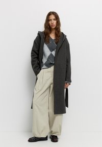 Grijze wollen jas met capuchon over een gestreept grijs-wit sweater, gecombineerd met losse beige broek en zwarte schoenen, staand tegen een effen achtergrond.