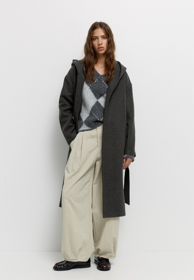 Grijze wollen jas met capuchon over een gestreept grijs-wit sweater, gecombineerd met losse beige broek en zwarte schoenen, staand tegen een effen achtergrond.