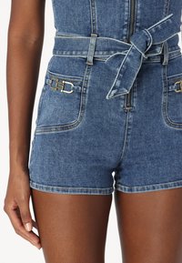 Närbild av blå denimshorts med framsida dragkedja, knytbart midjebälte och metallspännen på fickorna, som bärs av en person.