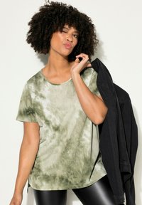 Camiseta verde tie-dye con mangas cortas, cuello redondo y una textura suave. Combinada con una chaqueta de mezclilla negra sobre un hombro.