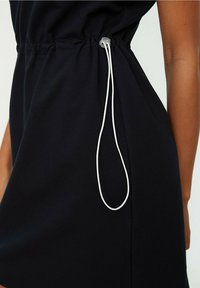 Czarna sukienka z marszczonym wcięciem w talii oraz białą regulowaną sznurówką po boku, noszona przez osobę z widoczną ręką.