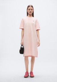 Ροζ φόρεμα με κοντά μανίκια, χαλαρή εφαρμογή και πλαϊνές σχισμές. Συνδυασμένο με ροζ παπούτσια και κρατώντας μια μικρή μαύρη τσάντα χειρός.