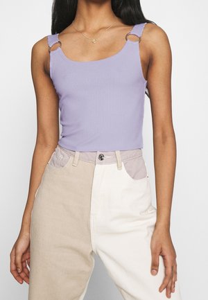 Femme portant un débardeur côtelé lavande avec des bretelles en anneau métallique et un pantalon taille haute bicolore beige et blanc, debout devant un fond uni.