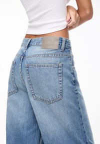 Jean en denim bleu clair avec taille haute, jambes larges et une pièce en cuir grise sur la ceinture arrière. Dispose de deux poches arrière.
