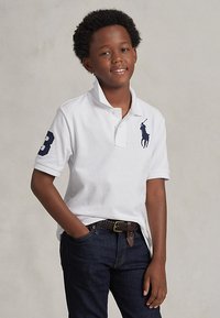 Lächelnder Junge mit lockigem Haar, der ein weißes Poloshirt mit einem dunkelblauen Logo und dunkelblaue Jeans trägt und mit einer Hand in der Tasche steht.