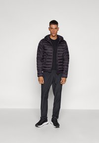 Giubbotto gonfiabile nero con cappuccio, design trapuntato, abbinato a una t-shirt nera e pantaloni scuri. Le scarpe presentano un accento di suola bianca.