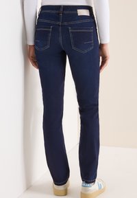 Jeans dżinsowe w ciemnoniebieskim kolorze, o dopasowanym kroju, z standardowymi kieszeniami z tyłu i widocznym przeszyciem, zestawione z białymi i niebieskimi sneakersami.