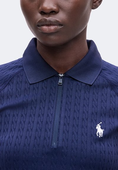 Gros plan d'une personne portant un polo bleu marine texturé avec fermeture éclair et un logo blanc brodé sur la poitrine gauche.