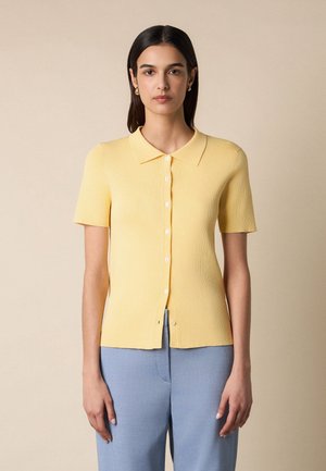 Donna con capelli scuri e lisci che indossa una camicia gialla a maniche corte con bottoni e pantaloni azzurri chiaro su sfondo beige.