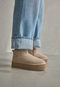 Beżowe zamszowe botki za kostkę z zaokrąglonym noskiem i lekko teksturowaną platformą, w zestawieniu z podwiniętymi jasnoniebieskimi dżinsami.