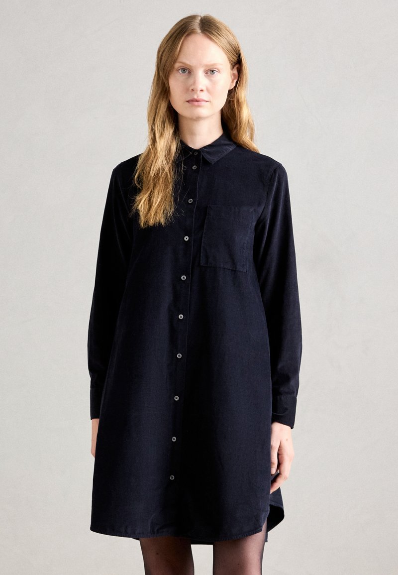 Donna con lunghi capelli biondi che indossa un vestito camicia nero a maniche lunghe con tasca sul petto, in piedi contro uno sfondo chiaro e uniforme.