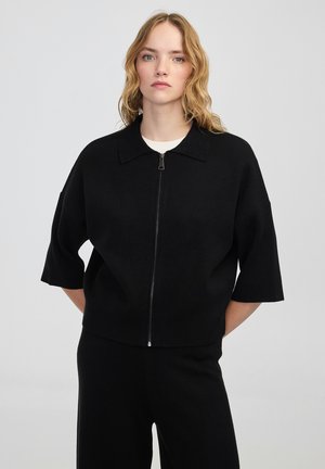 Donna con capelli biondi e mossi che indossa un maglione nero con zip e maniche larghe, abbinato a pantaloni neri, su uno sfondo semplice.