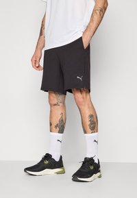 Zwarte sportshorts met zijsplitten, grijs logo, gecombineerd met zwarte sneakers met groene accenten en witte sokken. Eenvoudig wit T-shirt.
