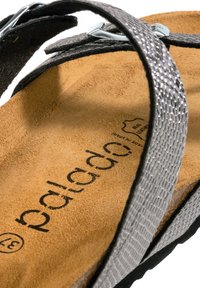 Sort sandal med en struktureret sølvrem, en brun ruskinds sål og en kontrasterende sort ydersål. Har en metalspænde detalje.