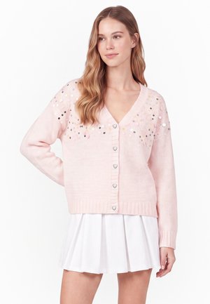 Junge Frau mit langen welligen Haaren, die eine hellrosa, mit Pailletten verzierte Strickjacke und einen weißen plissierten Rock trägt und mit einer Hand in der Hüfte steht.