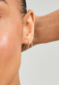 Boucles d'oreilles en or avec de petits cristaux transparents, présentant un design simple et courbé. Portées sur une oreille percée, mettant en avant un style minimaliste.