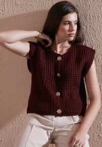Žena v texturovanej tmavoružovej bezrukávovej veste s zlatými gombíkmi, béžových nohaviciach, so zlatými náramkami a prsteňmi, pózuje pred texturovanou stenou.