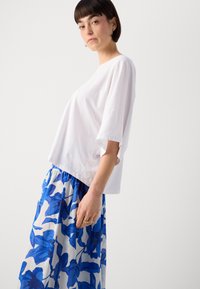 Haut blanc à manches courtes, ample, associé à une longue jupe bleue à motif floral avec une taille froncée. Texture lisse.