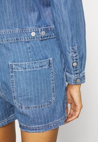 Vêtement en denim avec rayures verticales blanches, poche arrière, détails de bouton métallique et manche retroussée sur le bras et la main visibles d'une personne.