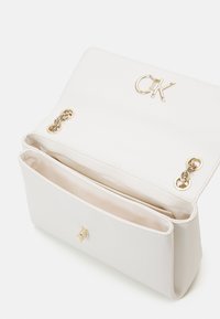 Bílá kožená kabelka se zlatým kováním, s magnetickým zavíráním na klopu a dvěma vnitřními přihrádkami. Vpředu reliéfní logo.