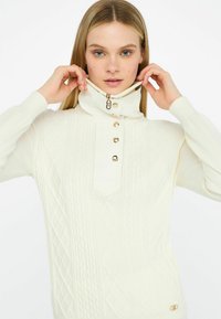 Pulover crem cu guler înalt, cu un model de tricotaj în cablu, accente cu nasturi aurii și detalii cu fermoar la guler. Textură moale și design ajustat.