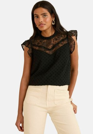 Black top featuring a layered lace yoke with a dotted texture, short ruffled sleeves, and a loose fit. Paired with light-colored pants.