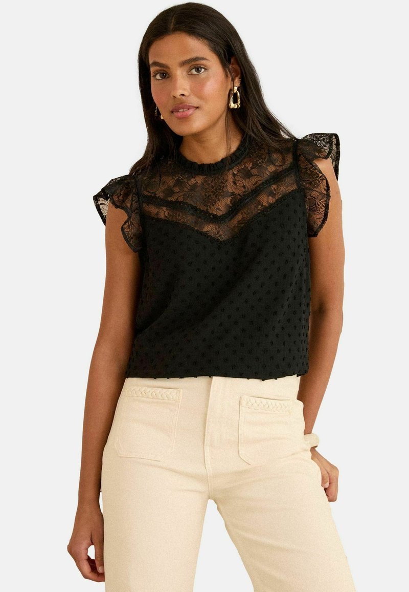 Top nero con un colletto in pizzo sovrapposto a strati con una texture a pois, maniche corte a rouche e una vestibilità ampia. Abbinato a pantaloni di colore chiaro.