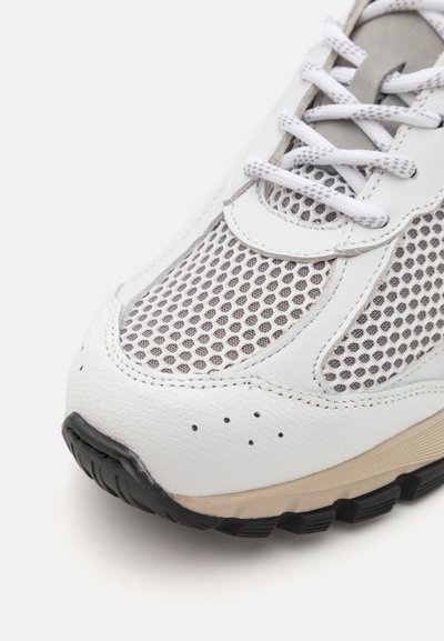 Baskets blanches en cuir avec des panneaux en maille grise, semelle extérieure en caoutchouc noir arrondi et lacets blancs plats.