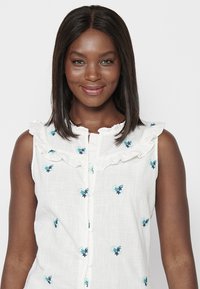 Weißes ärmelloses Bluse mit gerüschtem Kragen, ausgestattet mit blauen Blumenstickereien und einer Knopfverschluss vorne. Strukturierter Stoff.