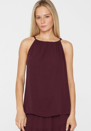 Femme blonde portant un haut dos-nu violet foncé, ample, debout devant un fond blanc uni.