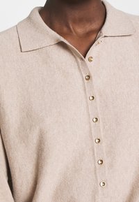 Polo in maglia beige con colletto e sette bottoni dorati sul davanti. Tessuto morbido con un motivo sottile.