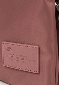 Recycled fabric bag in a muted pink tone. Features a textured surface and a rectangular patch with embossed lettering and logo.