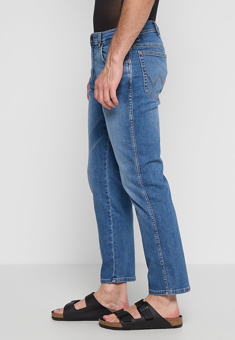 Osoba stoi bokiem, ubrana w niebieskie dżinsy i czarne sandały. Tło jest jednolite, białe, co podkreśla odzież.