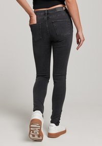 Jeans ajustados negros con cintura de media altura, que cuentan con bolsillos traseros estándar y una textura de denim suave, combinados con zapatillas blancas.