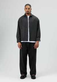 Donkergrijs wollen overhemd met knopen, losse pasvorm, gedragen over een wit T-shirt. Zwarte, loszittende broek en zwarte schoenen met gestructureerde veters.