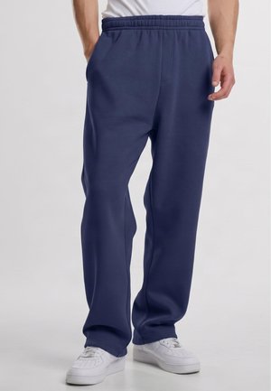 Homme portant un pantalon de jogging bleu marine à taille élastique et des baskets blanches, debout devant un fond clair uni.