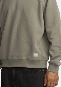 Olijfgroene katoenen sweatshirt met ribgebreide manchetten en onderrand, voorzien van een geweven label aan de onderzijde links. De stof voelt zacht aan.