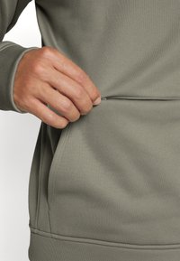 Main tirant la fermeture éclair sur la poche avant d'une veste de sport grise à manches longues.
